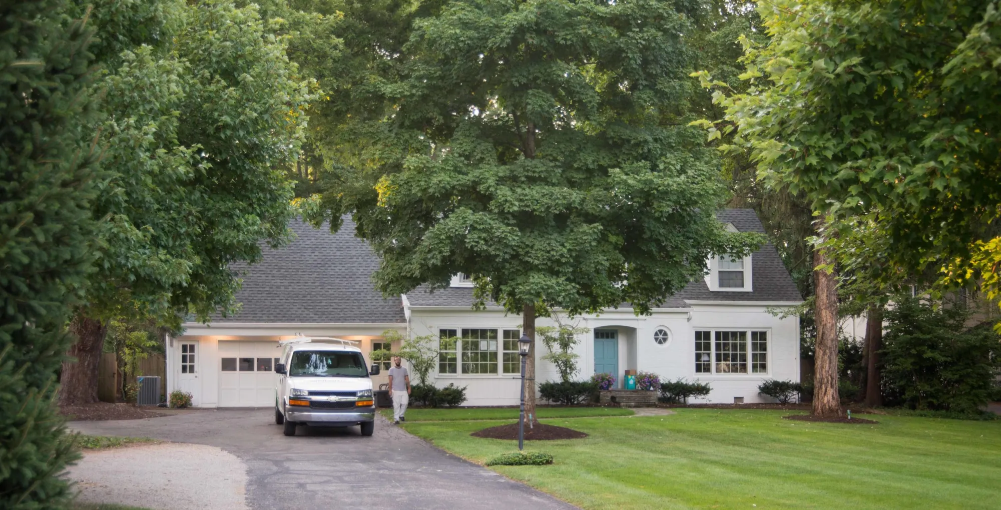 A white house with a gray roof under tall shade trees, an ALL IN van parked in the driveway